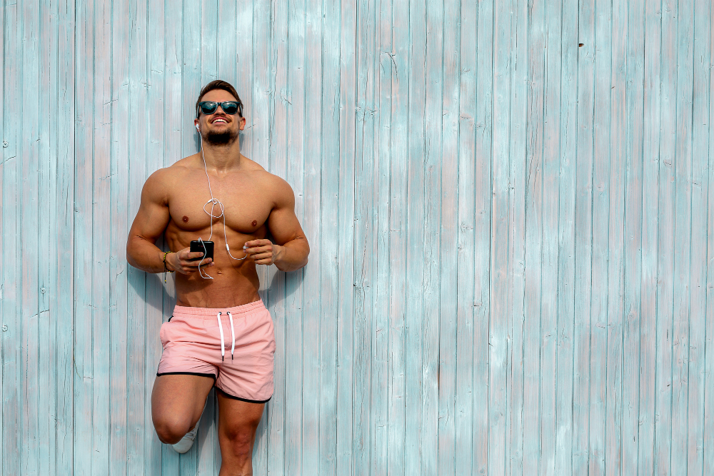 Umięśniony mężczyzna w różowych kąpielówkach, słuchający muzyki, na tle z desek pomalowanych na niebiesko. 