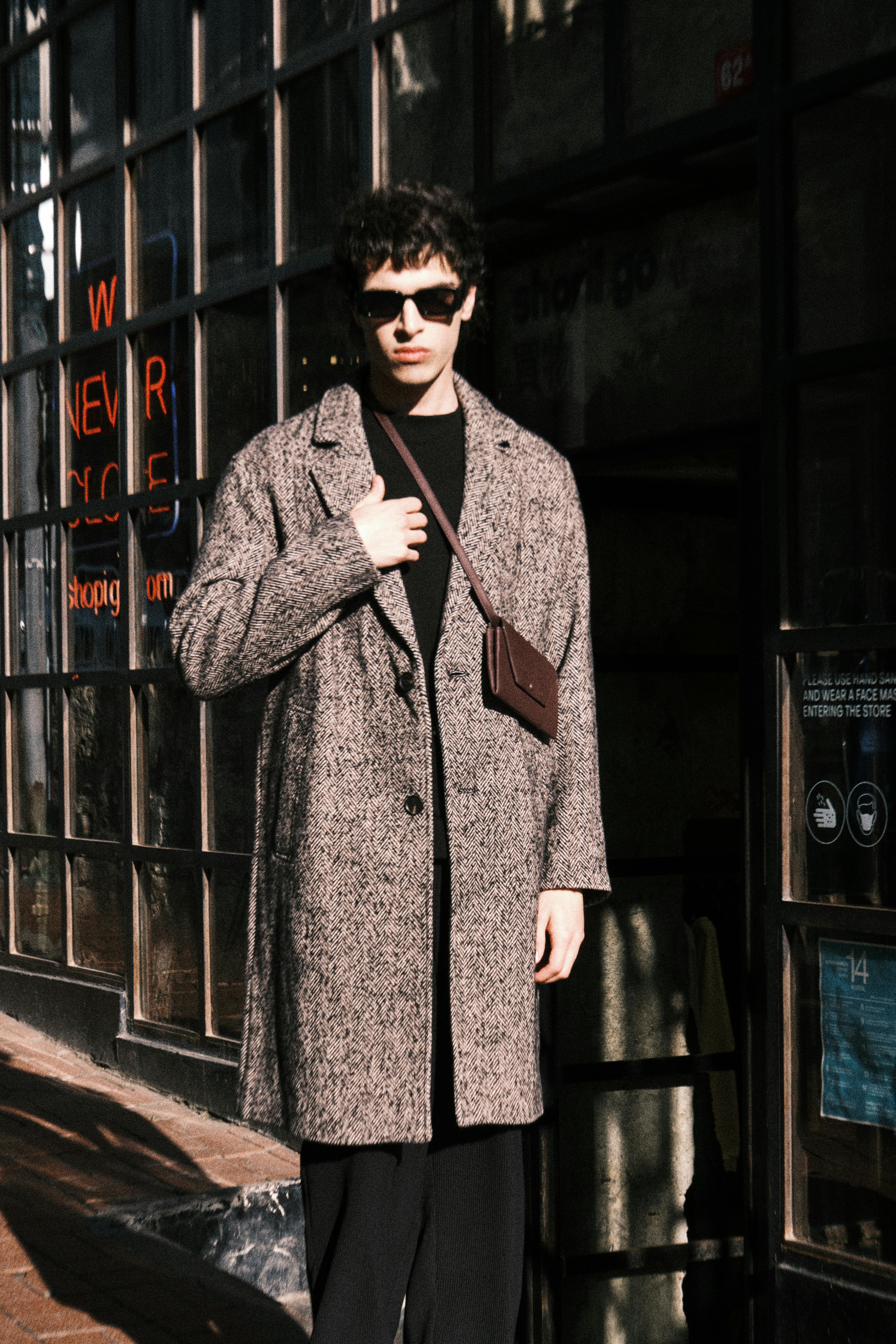 Mężczyzna w stylizacji na zimę z płaszczem, bluzą, okularami przeciwsłonecznymi oraz portmonetką.